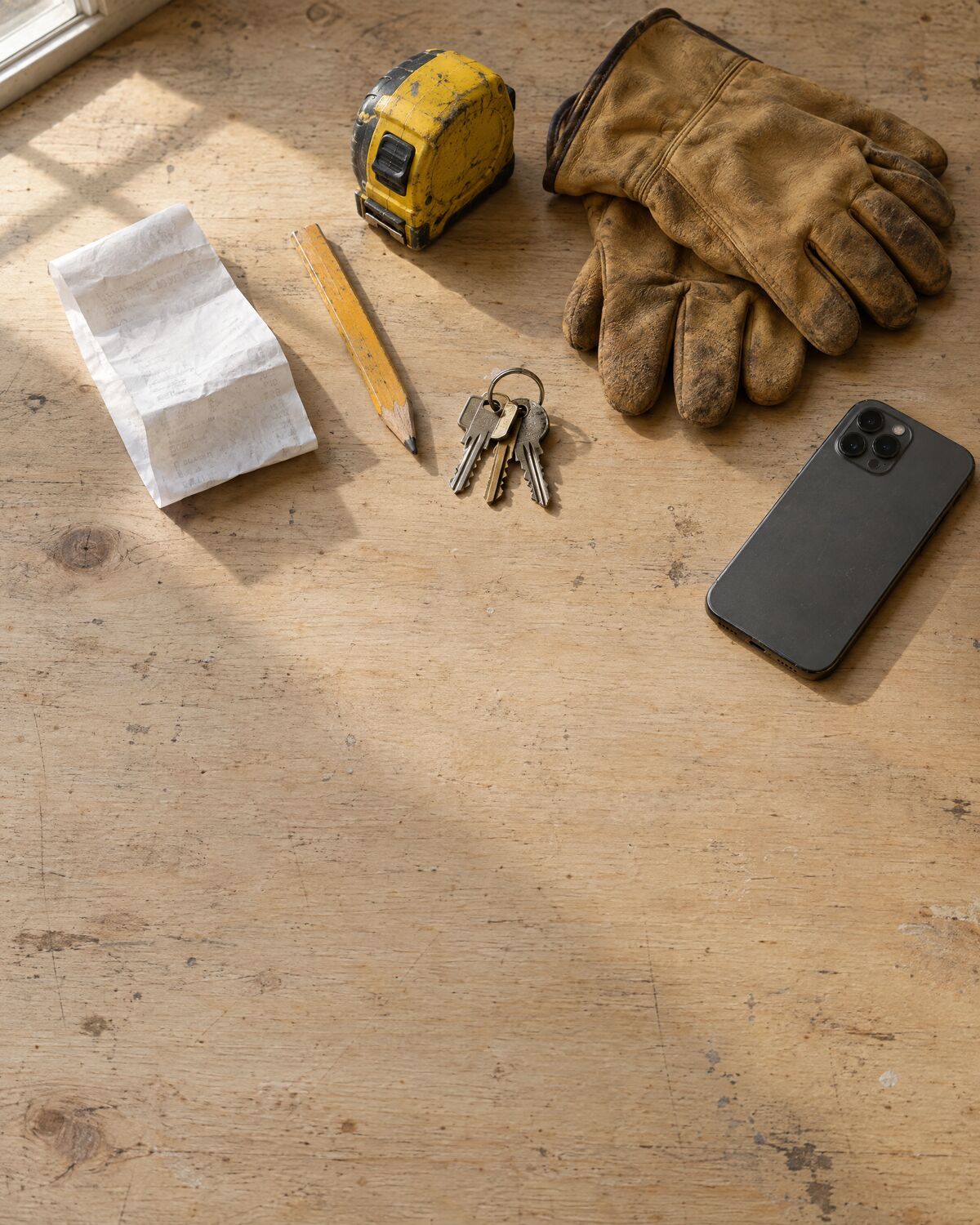 A receipt, a pencil, a tape measure, keys, work gloves and an iPhone lying face down on a workbench